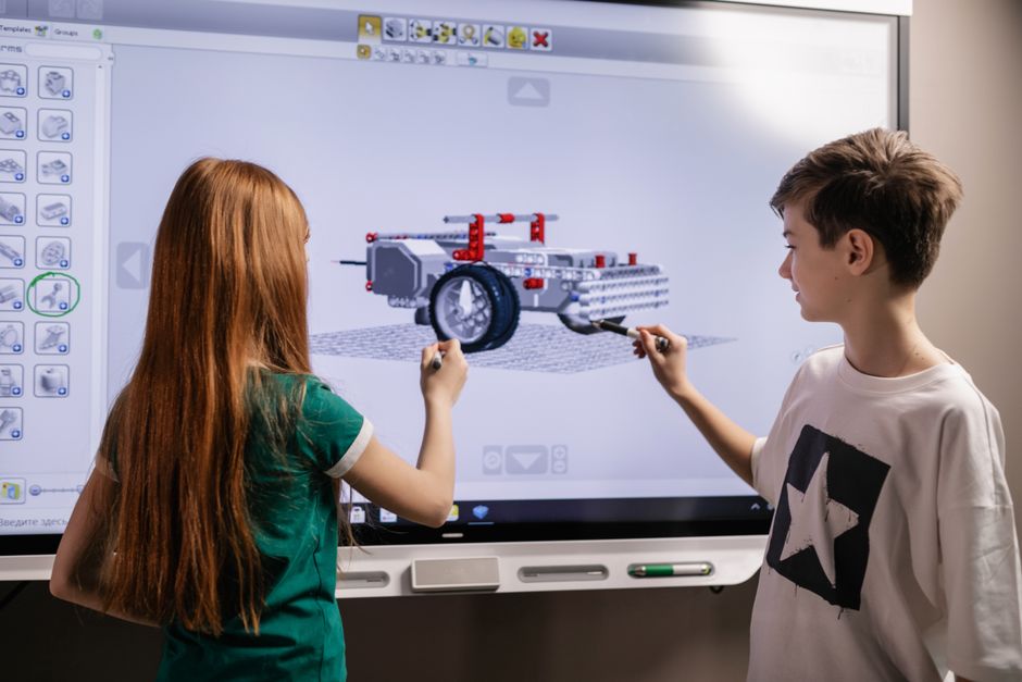 Duplo de crianças interagindo com um projeto de modelagem 3D de um dispositivo tecnológico em uma lousa digital, demonstrando inovação e aprendizado. 