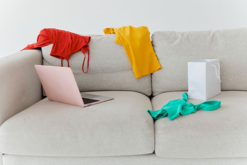 Cama desarrumada com roupas, laptop e embalagem, retratando o caos doméstico e a rotina diária.