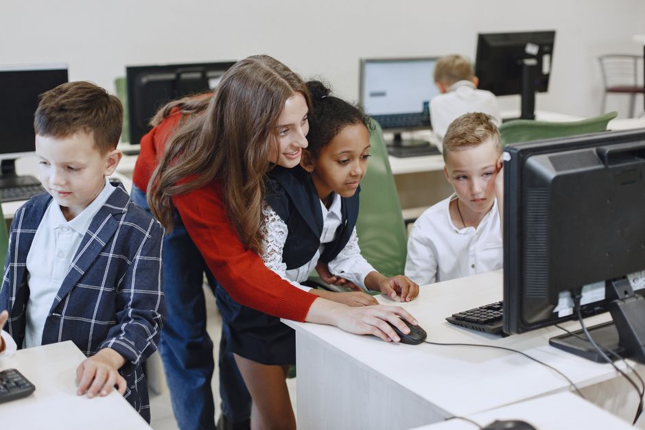 Professora orientando crianças em aula de informática, promovendo aprendizado tecnológico e colaboração.