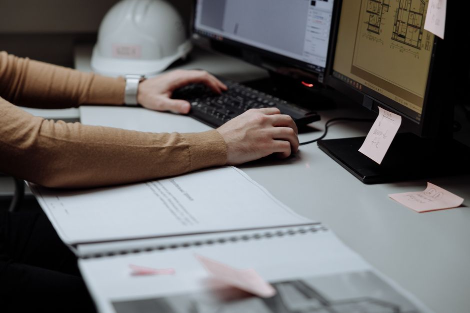 Pessoa trabalhando com projetos de arquitetura e design em ambiente de escritório, utilizando computador e documentos técnicos.