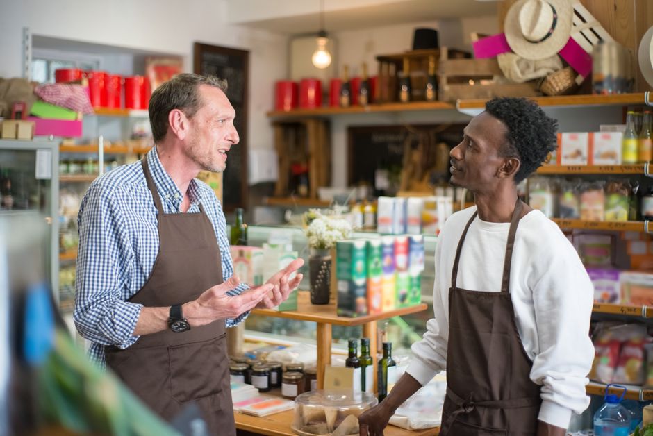 Dois comerciantes conversando em uma loja de produtos naturais ou alimentos artesanais, demonstrando interação e atendimento ao cliente.