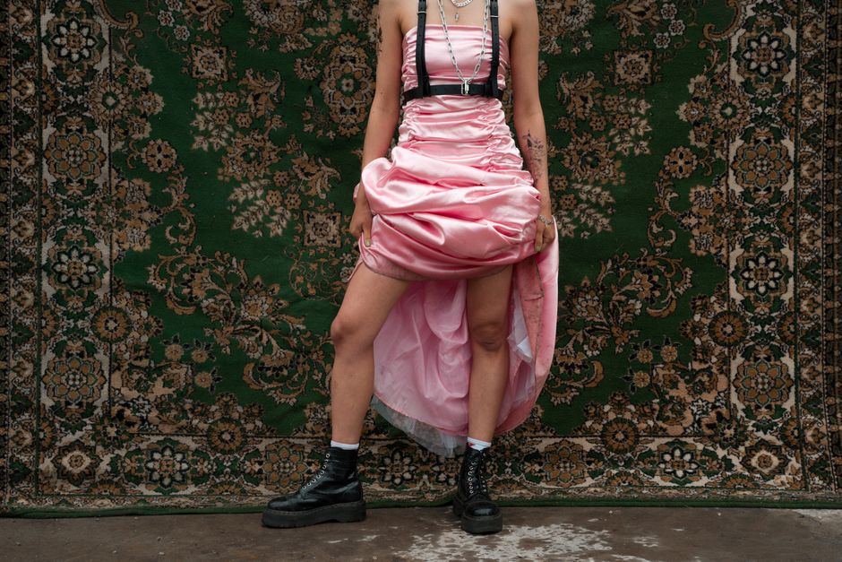 Pessoa usando vestido rosa de festa com postura confiante sobre um tapete decorativo, transmitindo uma expressão de individualidade e estilo.