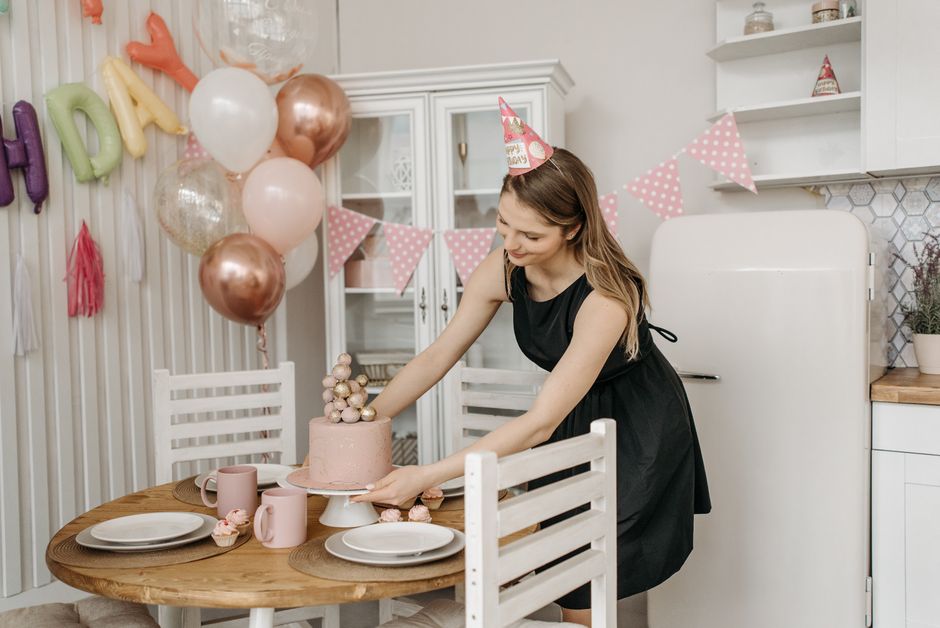 Mulher decorando uma mesa de festa de aniversário com tema feminino, usando chapéu de festa e ambiente festivo.