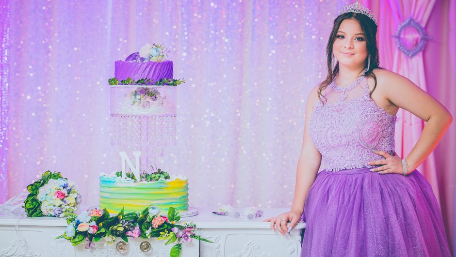 Juíza usando traje formal e tiara, comemorando uma ocasião especial ao lado de um bolo decorado, em um ambiente festivo com fundo rosa e brilho.