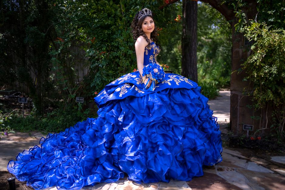 Jovem usando vestido de festa azul com detalhes dourados em um cenário ao ar livre, sugerindo elegância para eventos sociais ou ensaio fotográfico.