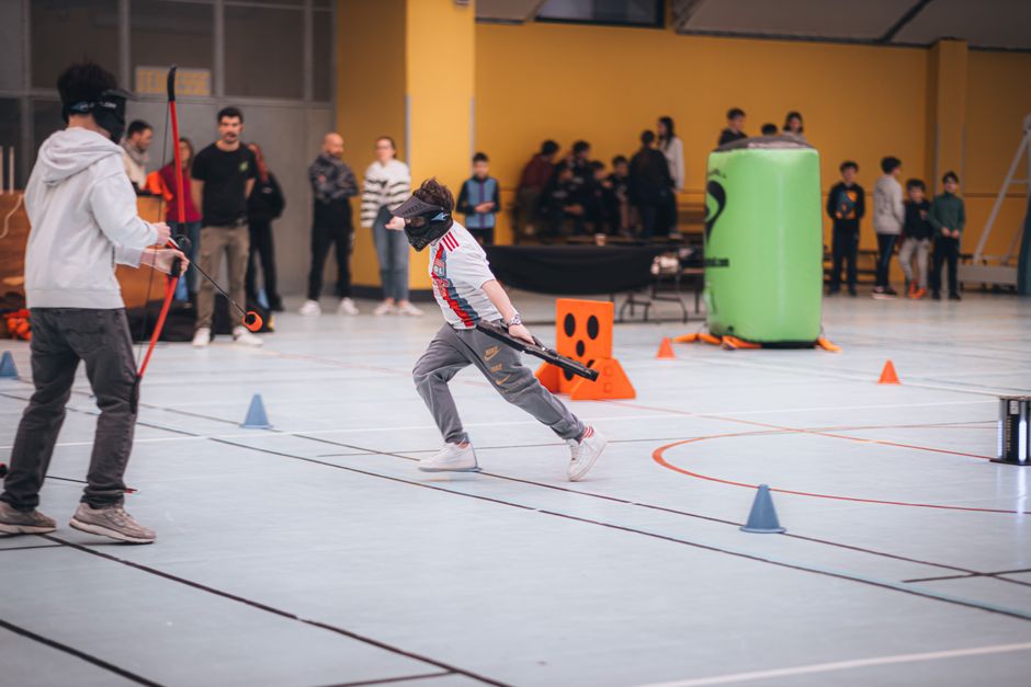 Imagem de uma criança participando de um jogo de tiro com arco em um evento indoor, com várias pessoas observando ao fundo, sob uma parede amarela.