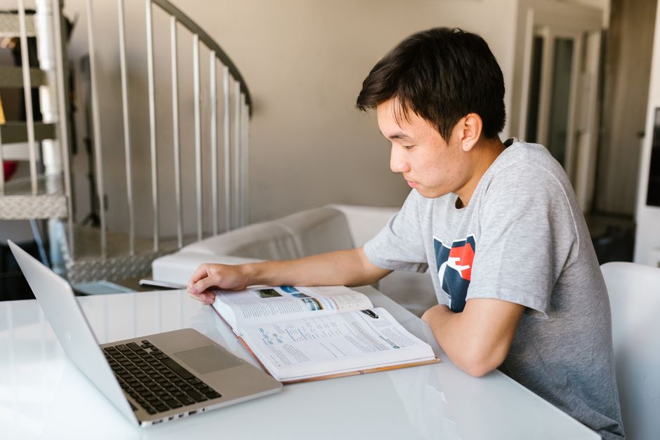 Jovem estudando com foco e concentração em um ambiente doméstico, usando laptop e livros abertos ao redor.