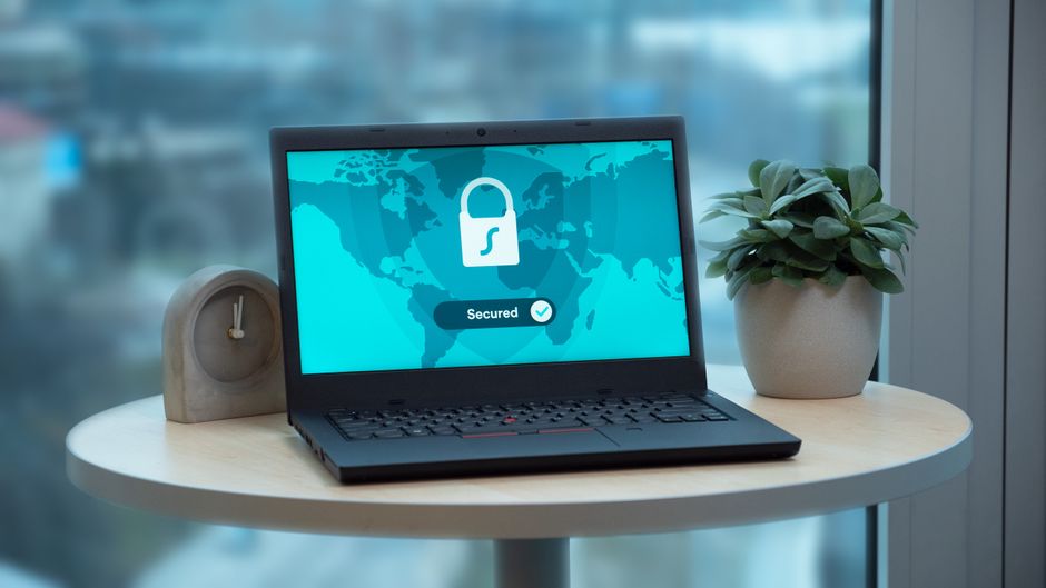 Laptop exibindo mensagem de segurança digital em um ambiente de trabalho moderno, com planta e relógio ao lado, próxima a janelas com vista urbana.