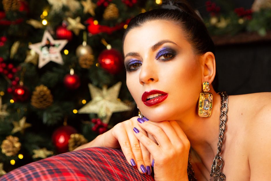 Mulher com maquiagem marcante e acessórios elegantes posando em frente a uma árvore de Natal decorada, transmitindo sofisticação e o espírito festivo.