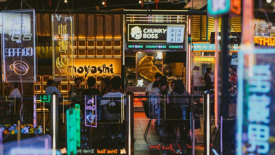 Ambiente de uma loja de fast food com iluminação neon vibrante e pessoas apreciando suas refeições, criando uma atmosfera moderna e movimentada.