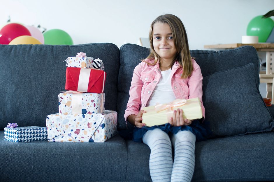 Menina sorridente sentada em um sofá, segurando presentes de aniversário ao lado de uma pilha de embrulhos coloridos.