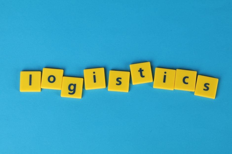 Imagem com blocos amarelos formando a palavra 'Logistics' sobre fundo azul, representando a gestão de logística ou transporte.