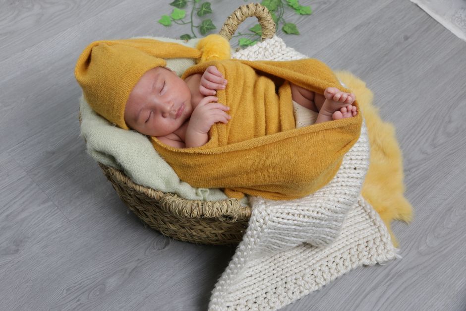 Bebê dormindo aconchegado em uma cesta decorada, vestindo roupinha amarela e cobertura de lã, em ambiente acolhedor.