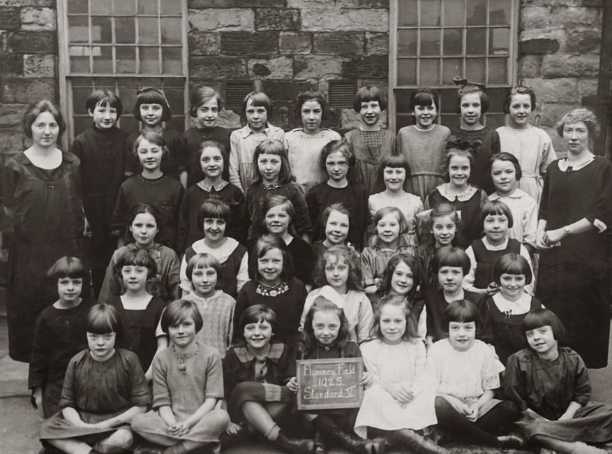 Grupo de crianças em aula escolar vintage, sorrindo para a câmera em frente a uma parede de tijolos, com uma professora ao lado