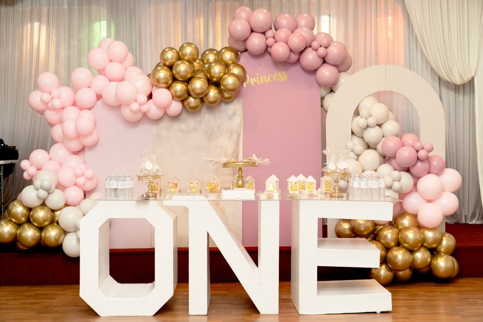 Decoração de festa infantil com tema princesa, destaque para balões rosas, dourados e brancos ao redor de um painel com a palavra 'PRINCESS'.