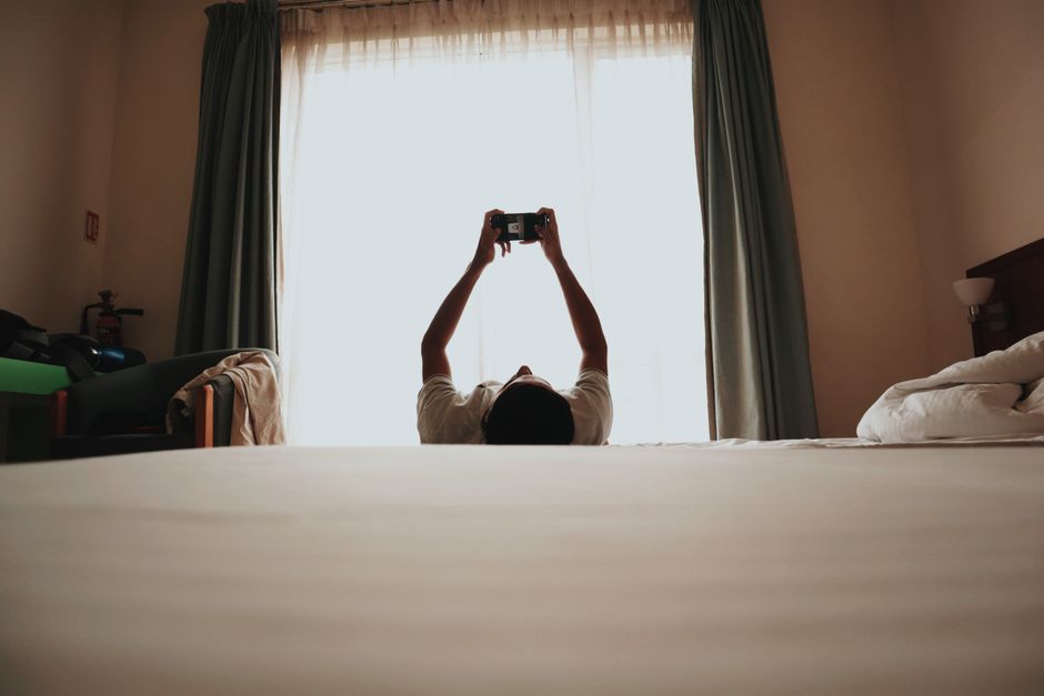 Pessoa tirando uma selfie deitada na cama em um quarto com cortinas de cortina clara, com foco na atmosfera de relaxamento e rotina diária.