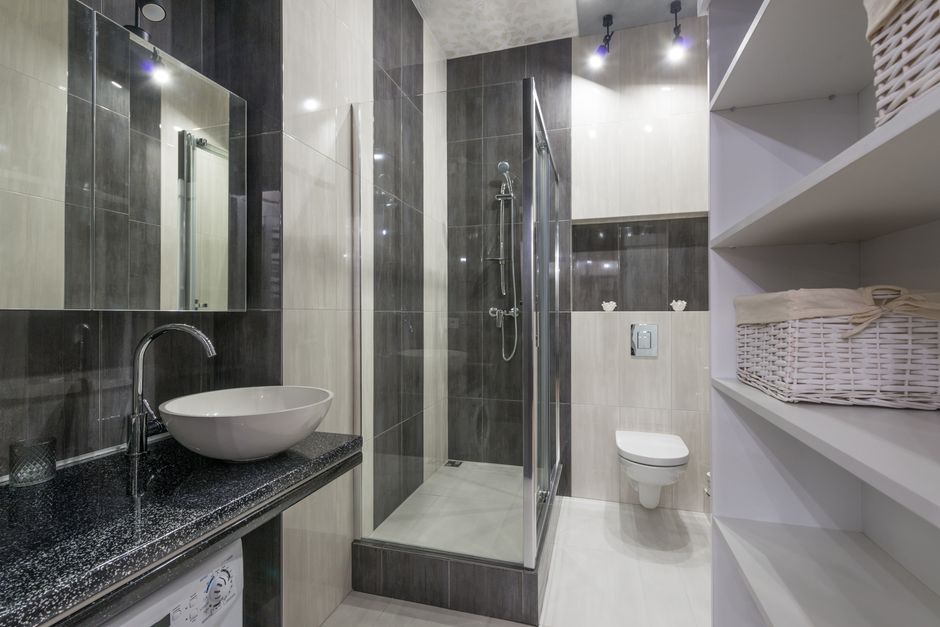 Imagem de um banheiro moderno com acabamento em tons de cinza e detalhes em preto, destacando a área de banho, vaso sanitário e prateleiras de armazenamento.