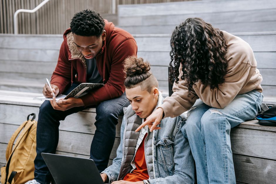 Grupo de jovens estudantes discutindo projetos acadêmicos ao ar livre, com notebooks e materiais de estudo.