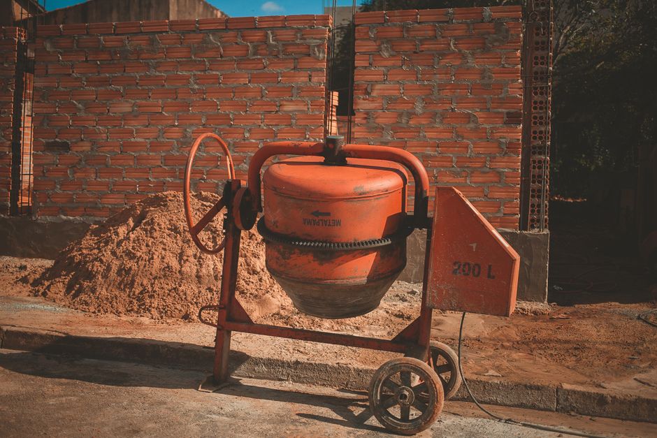 Imagem de um misturador de concreto em um canteiro de obras com parede de tijolos ao fundo, destacando o processo de construção civil.
