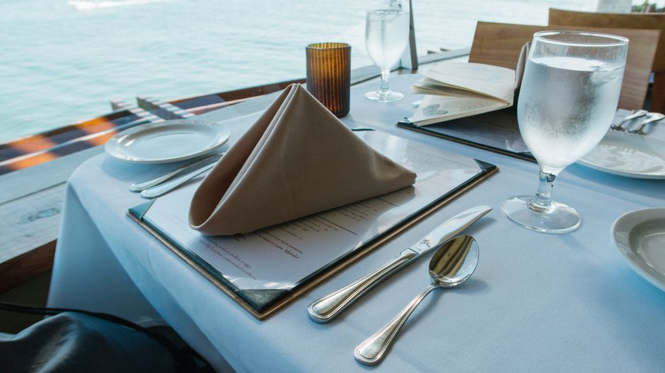 Mesa de restaurante elegantemente arrumada com detalhes de serviço, ao lado de um cenário marítimo relaxante.