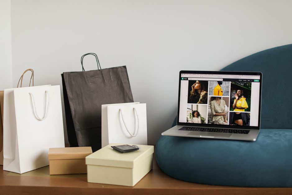 Imagem que ilustra o conceito de compras online de moda, com laptop exibindo fotos de roupas e várias sacolas de compras ao lado.