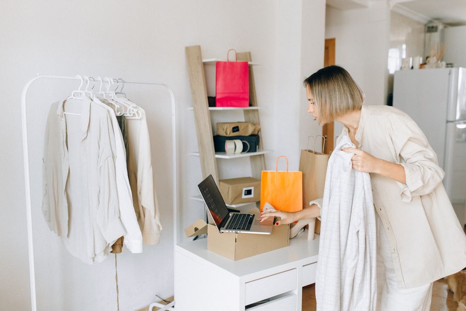 Mulher fazendo compras online enquanto organiza roupas e utensílios domésticos em casa.