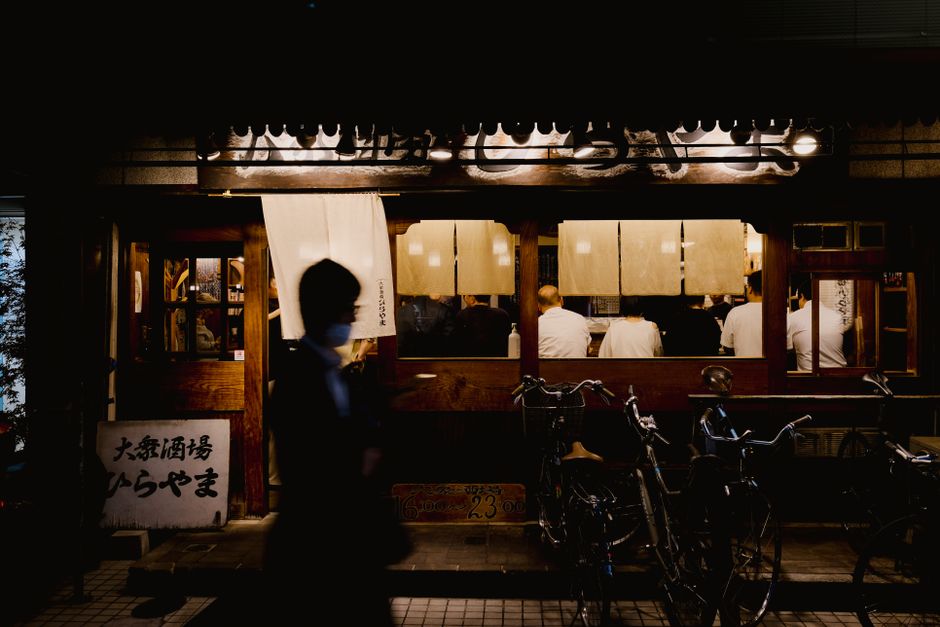 Foto de um bar tradicional japonês à noite, com clientes apreciando suas refeições e bebidas em um ambiente acolhedor.