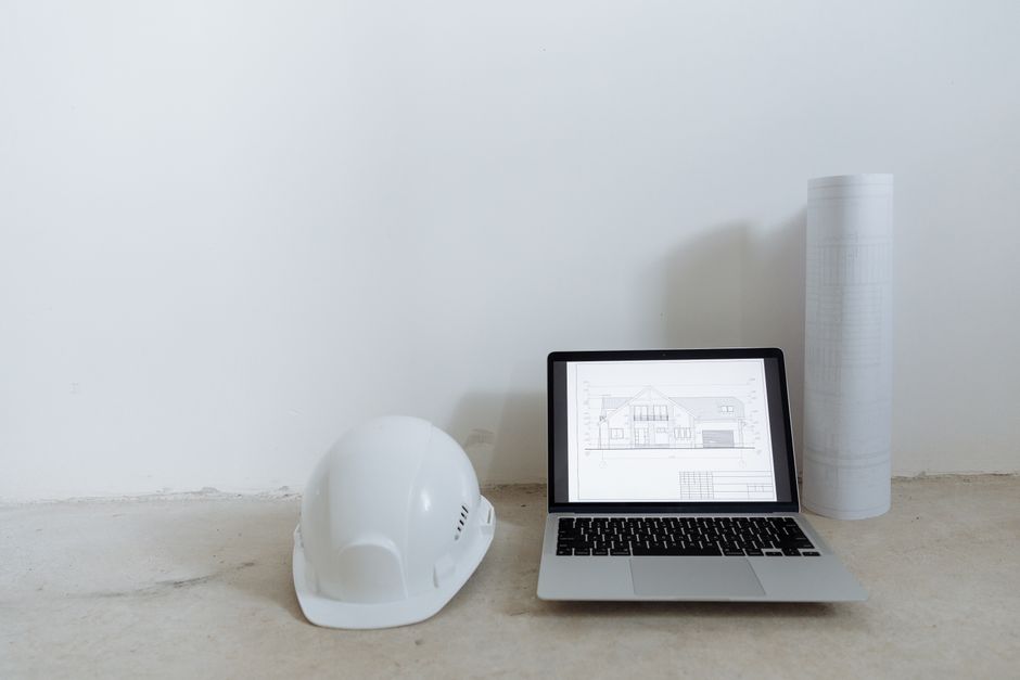 Imagem de um ambiente de trabalho com um capacete de segurança, um laptop exibindo um projeto arquitetônico e rolos de plantas, representando planejamento e gestão de projetos de construção civil.
