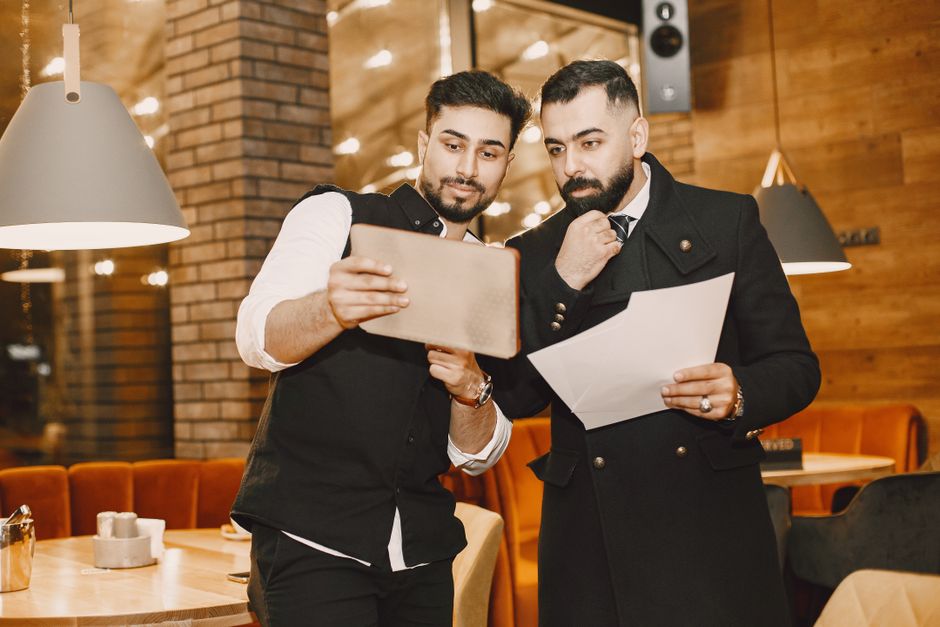 Dois homens conversando e analisando informações em um ambiente de restaurante, sugerindo uma discussão ou planejamento de negócios informal.