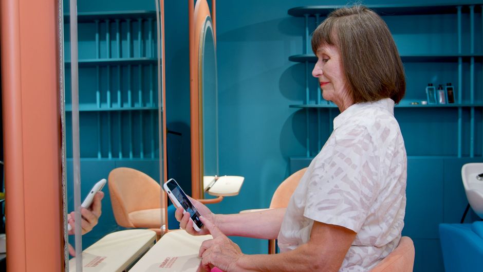 Mulher idosa usando smartphone em ambiente moderno de beleza ou estética, com foco na tecnologia aplicada a cuidados pessoais.