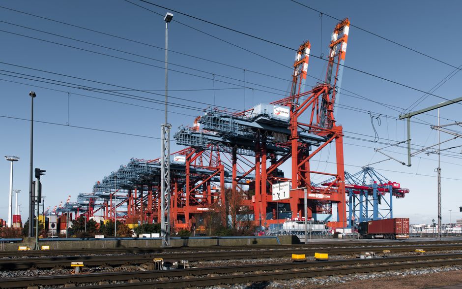 Imagem de um terminal portuário com guindastes de carga e trilhos ferroviários, representando logística e transporte de cargas.