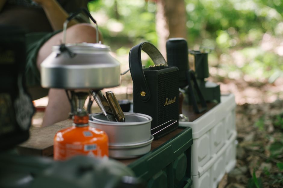 Imagem que mostra um equipamento de camping, incluindo uma geladeira portátil, um fogareiro e utensílios, em um ambiente ao ar livre rodeado de árvores.