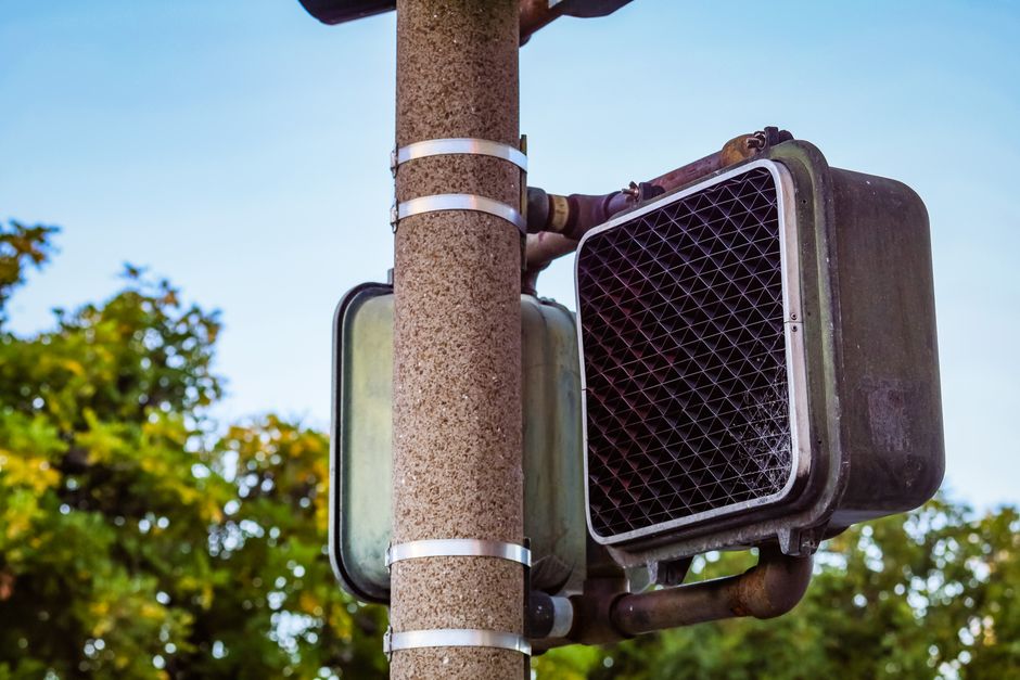 Imagem de um radar ou dispositivo de fiscalização instalado em poste para controle de velocidade, sobre céu claro e árvores ao fundo.