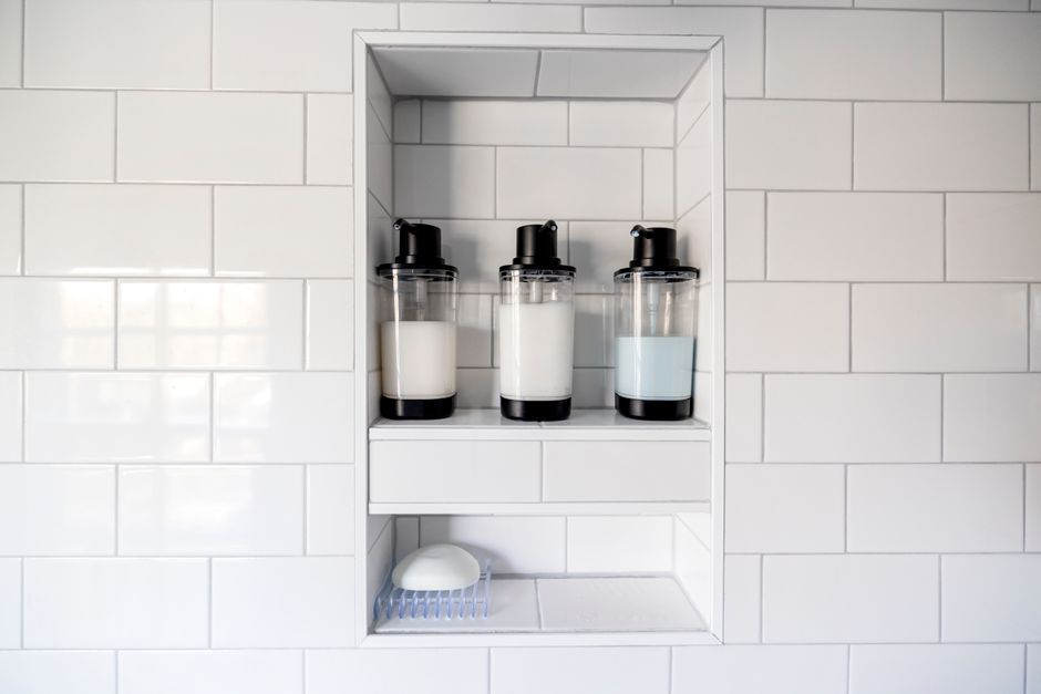 Imagem de uma prateleira embutida em um revestimento de azulejos brancos, com potes de sabonete líquido ou loção em dispensers transparentes, sugerindo um ambiente de banheiro ou lavabo bem organizado.