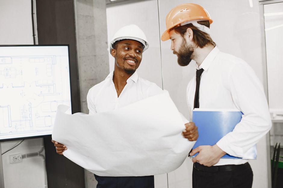 Engenharia civil ou arquitetura em discussão durante uma reunião de projeto, com dois profissionais usando capacetes de segurança e analisando um plano.