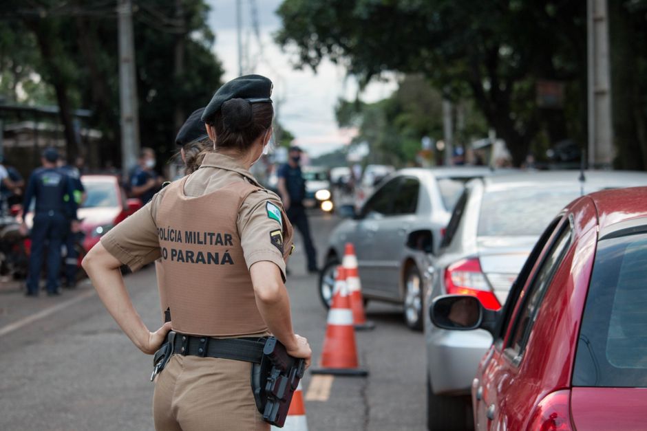 Policial Militar do Paraná realizando controle de trânsito em uma rua movimentada com vários carros e cones de sinalização ao fundo.