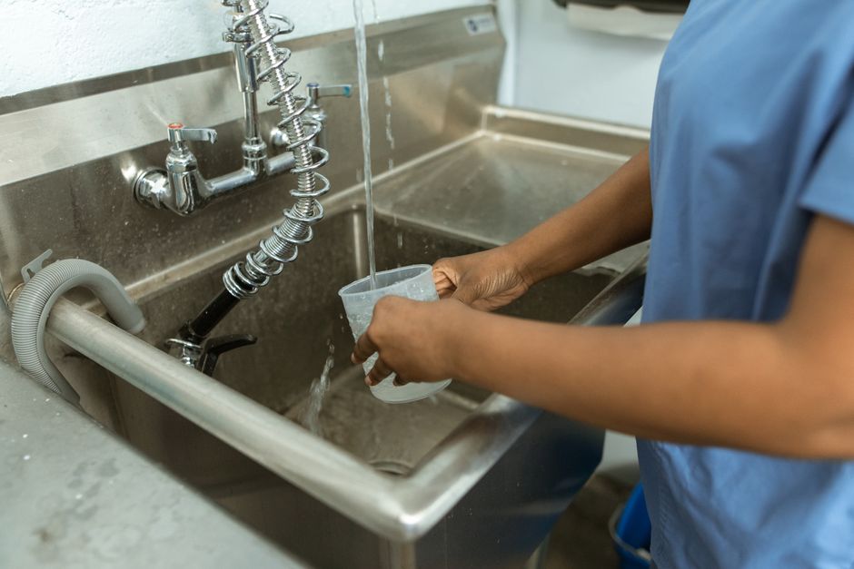 Imagem de uma pessoa lavando um copo sob torneira de uma cozinha ou cozinha industrial, ressaltando a higiene e preparo de alimentos ou bebidas.