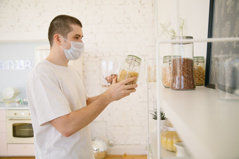 Pessoa com máscara analisando alimentos em potes de vidro, em ambiente de cozinha ou loja de alimentos, com foco na apresentação e armazenamento de produtos alimentícios.