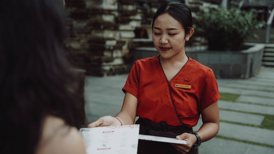 Uma funcionária de hotel sorridente entregando o menu ou o cartão de serviço para um cliente ao ar livre, demonstrando hospitalidade e atendimento ao cliente.