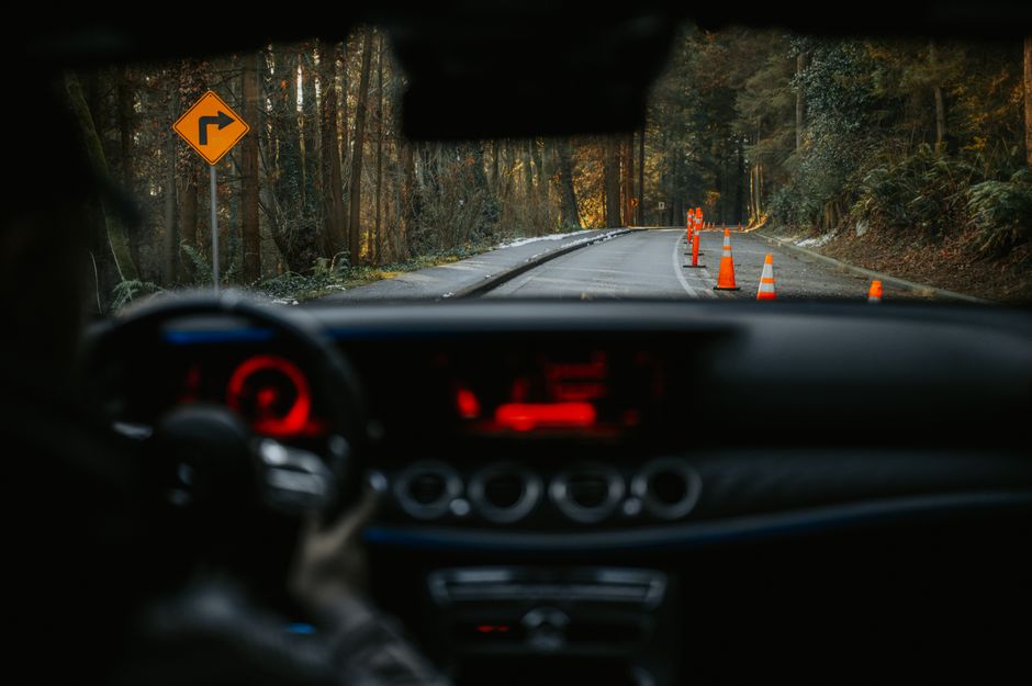 Imagem de uma estrada sinuosa vista do interior de um carro, com cones de trânsito e sinalização de curva à direita, cercada por uma floresta durante o dia.