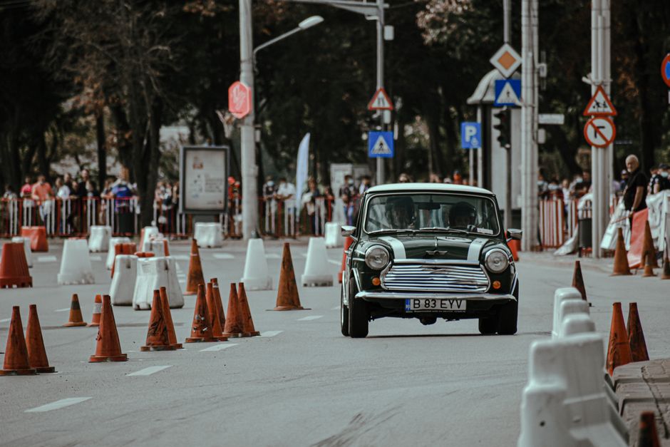 Carro clássico em uma rua fechada ao trânsito, com cones de segurança e espectadores ao fundo, em um evento urbano de carros antigos.
