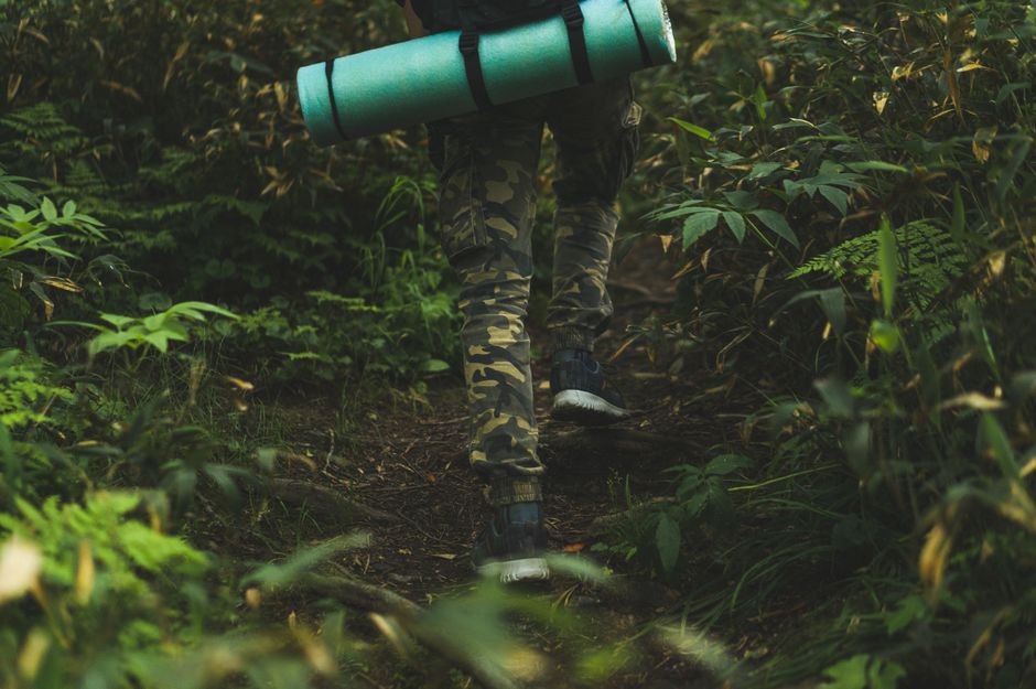 Pessoa caminhando por uma trilha na floresta com uma esteira de dormir nas costas, em uma aventura ao ar livre.