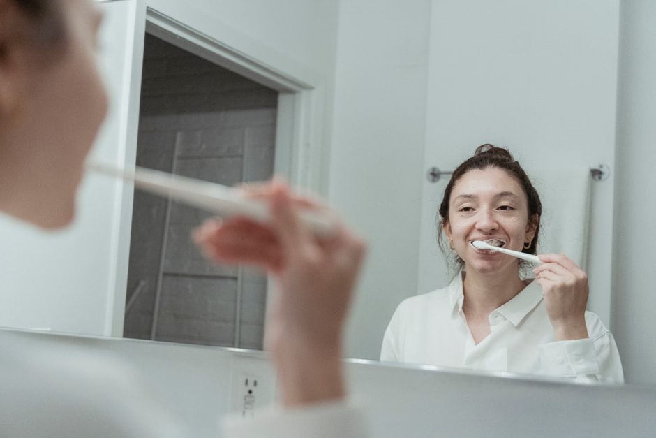 Mulher escovando os dentes no banheiro, refletida no espelho, em uma rotina de cuidado pessoal matinal.