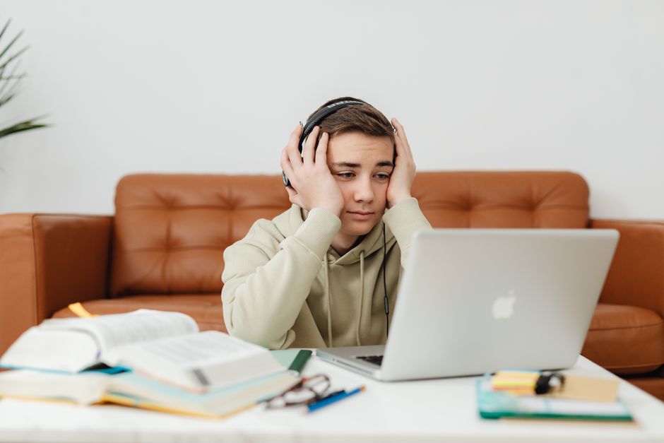 Jovem estudando e enfrentando dificuldades com tarefas escolares ou acadêmicas enquanto usa um laptop e fones de ouvido em um ambiente doméstico.