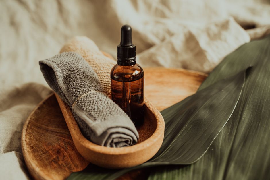 Imagem de produtos para cuidado pessoal e bem-estar, incluindo uma toalha de rosto, uma garrafinha de óleo essencial e folhas de plantas, sugerindo um ambiente de spa ou rotina de autocuidado.