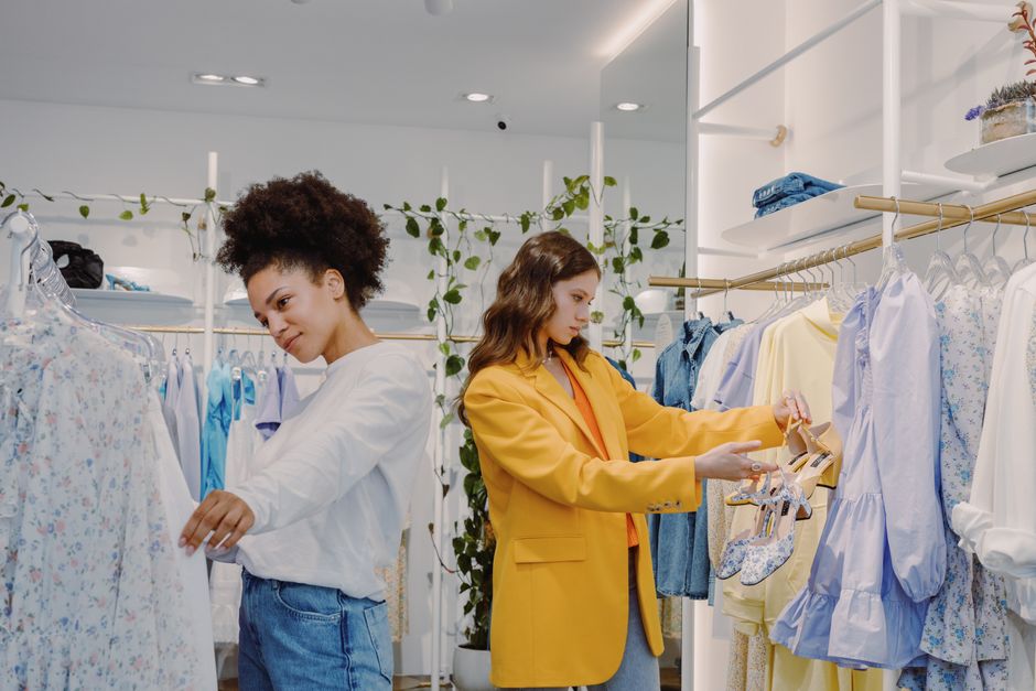 Duas mulheres fazendo compras em uma loja de roupas, uma analisando uma peça e a outra escolhendo sapatos, representando tendências de moda e consumo.