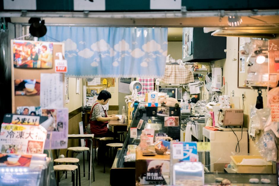 Cena de uma pequena loja ou restaurante japonês com uma mulher trabalhando no atendimento, ambiente aconchegante e decorado com itens diversos.
