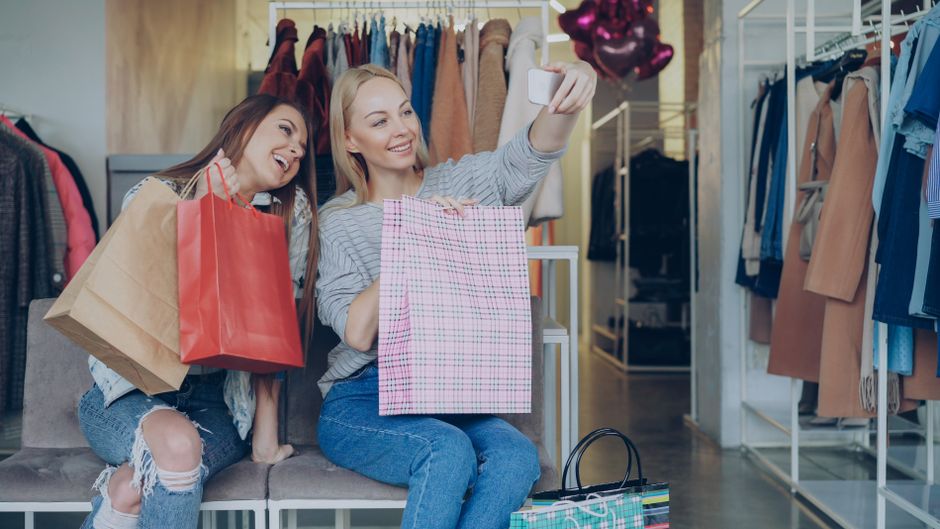 Duas mulheres felizes tirando selfie enquanto fazem compras de roupas em uma loja de moda feminina.