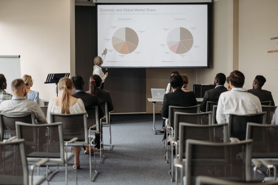 Professora explicando dados de mercado em uma sala de treinamento corporativo para uma audiência de profissionais de negócios.