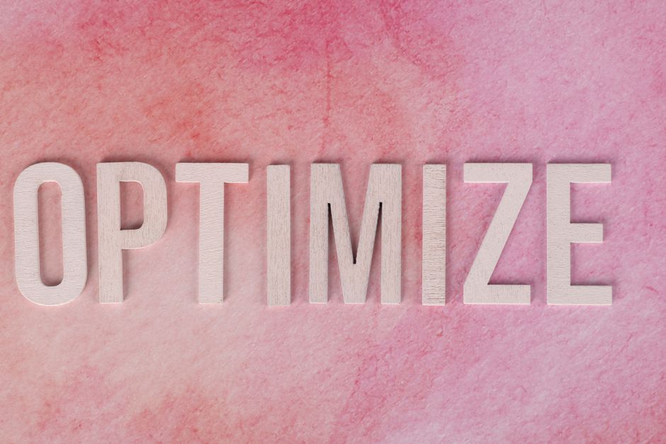 Imagem com letras de madeira formando a palavra 'OPTIMIZE' sobre fundo rosa, representando o tema de eficiência e melhorias em processos ou negócios.